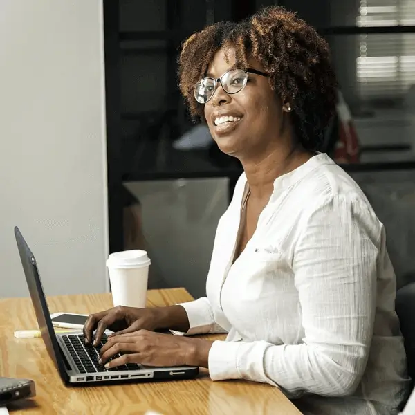 Intentional Approach to Mentorship. Woman typing on laptop at a desk with a smile on her face and a cup of coffee next to her 