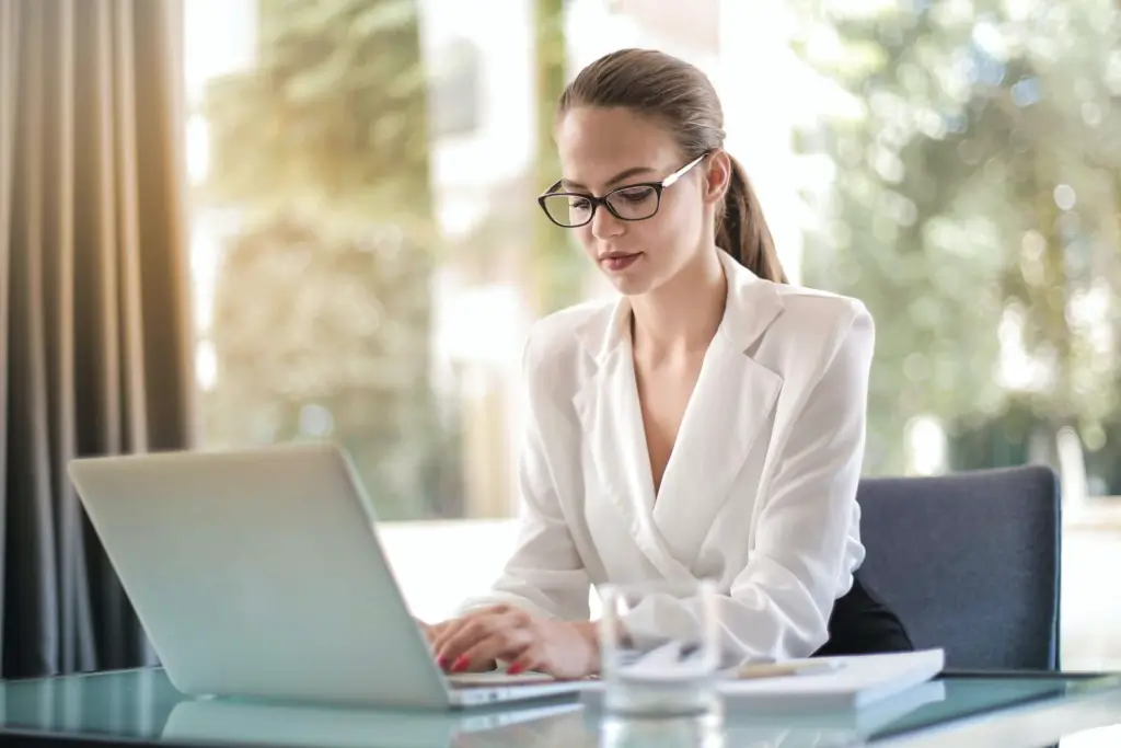 A person wearing glasses and a white blazer works on a laptop at a desk in a bright indoor space.