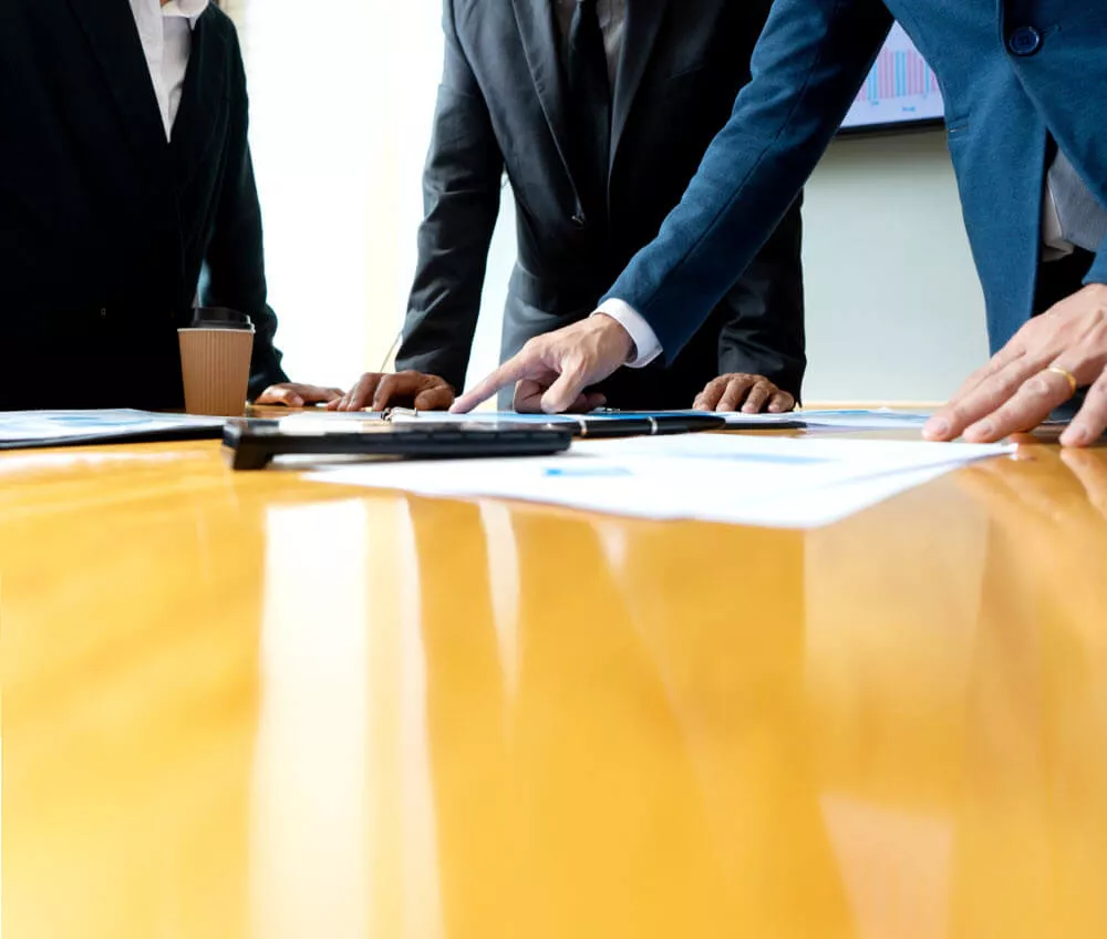 three people in business suits leaning over a table with paper, pens, and a calculator. one of them is pointing at a document.