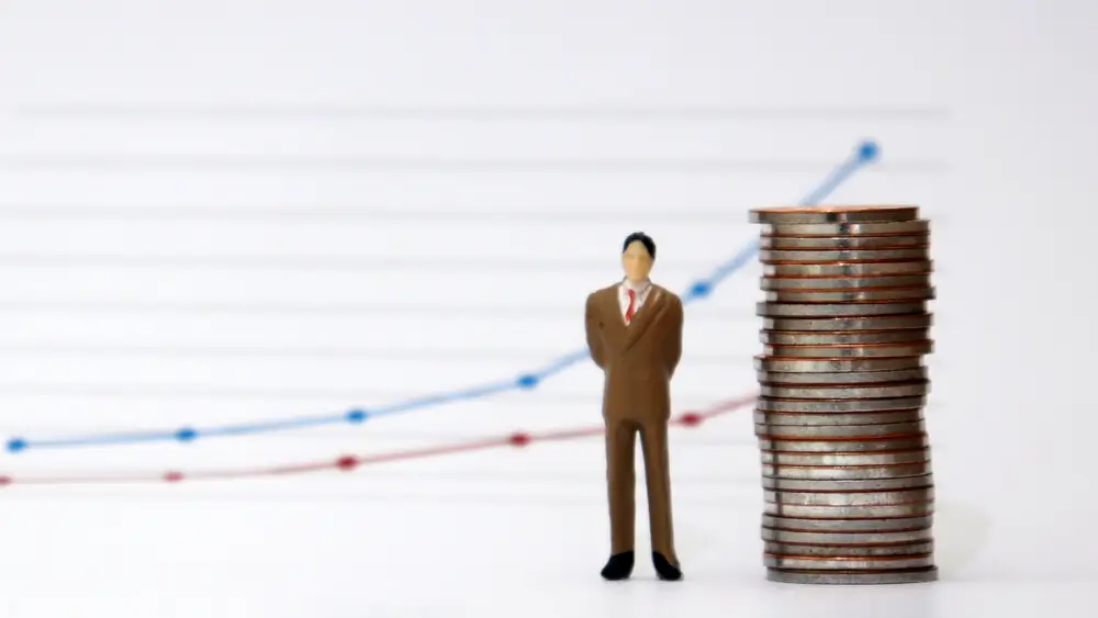 Photo of a miniature toy man in a business suit standing next to a stack of coins. A blurry line graph can be seen in the background.