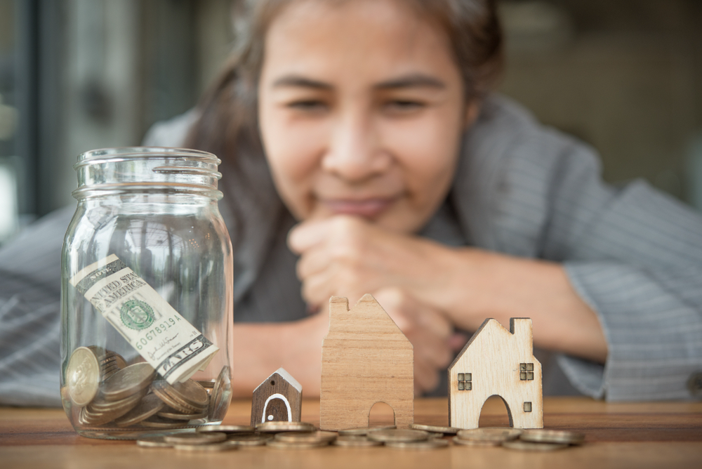 Photo of a woman smiling thoughtfully at a jar full of coins and three toy houses. She took our Personal Finance series, and now she's saving up to buy a house.