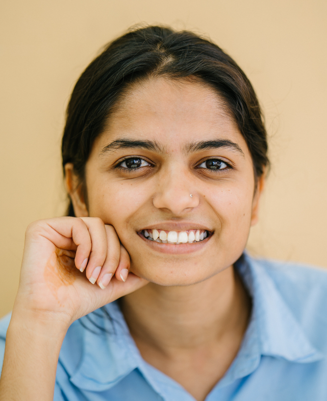 Smiling woman with dark hair pulled back, wearing a light blue button-up shirt, resting her chin on her hand against a neutral beige background.