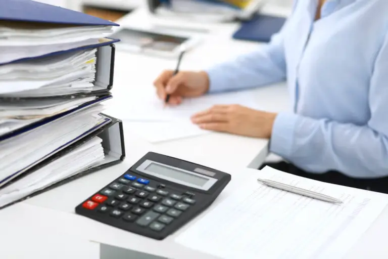 Person working at a desk with stacks of binders, financial documents, and a calculator, reviewing and recording accounting data.