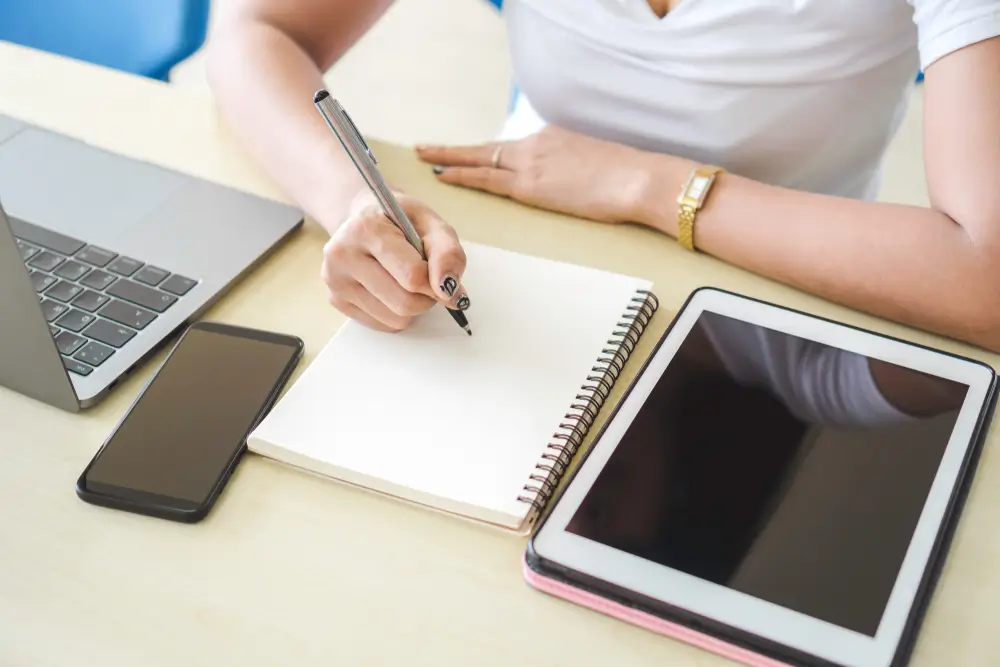 A student taking notes on paper with an iPad, smartphone, and laptop beside her.