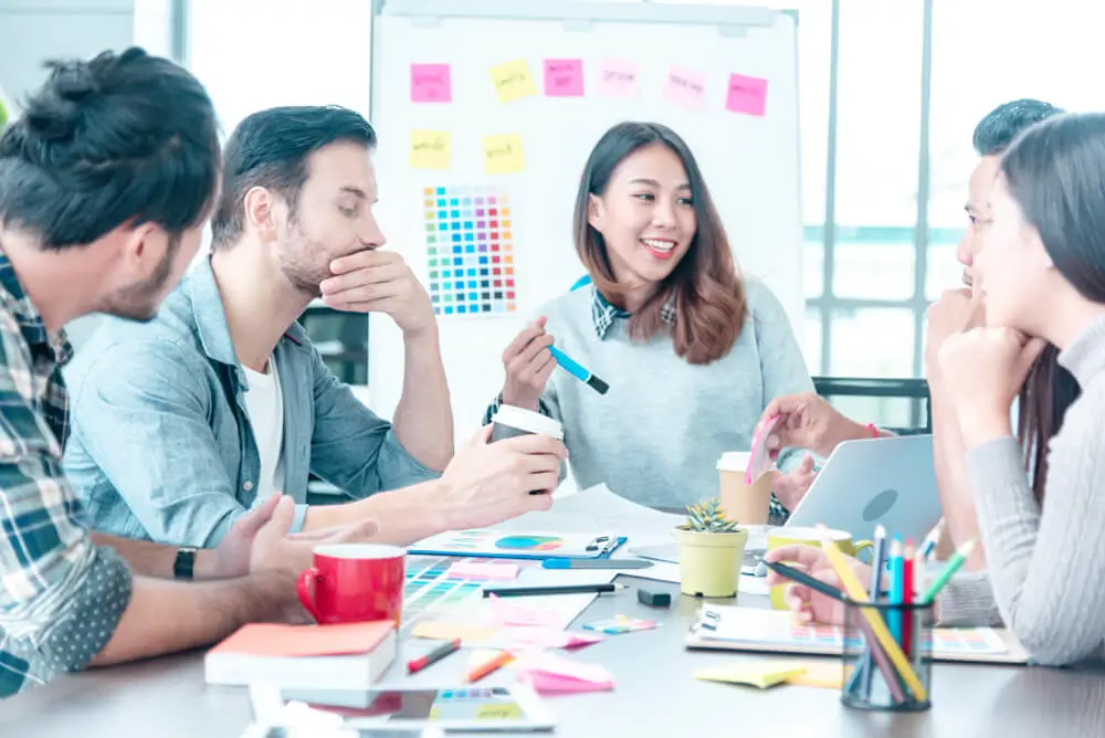 A group of five young professionals having a creative discussion around a table with color swatches, notebooks, and laptops, in a bright modern office. One woman is smiling while holding color samples, and others are engaged in conversation.