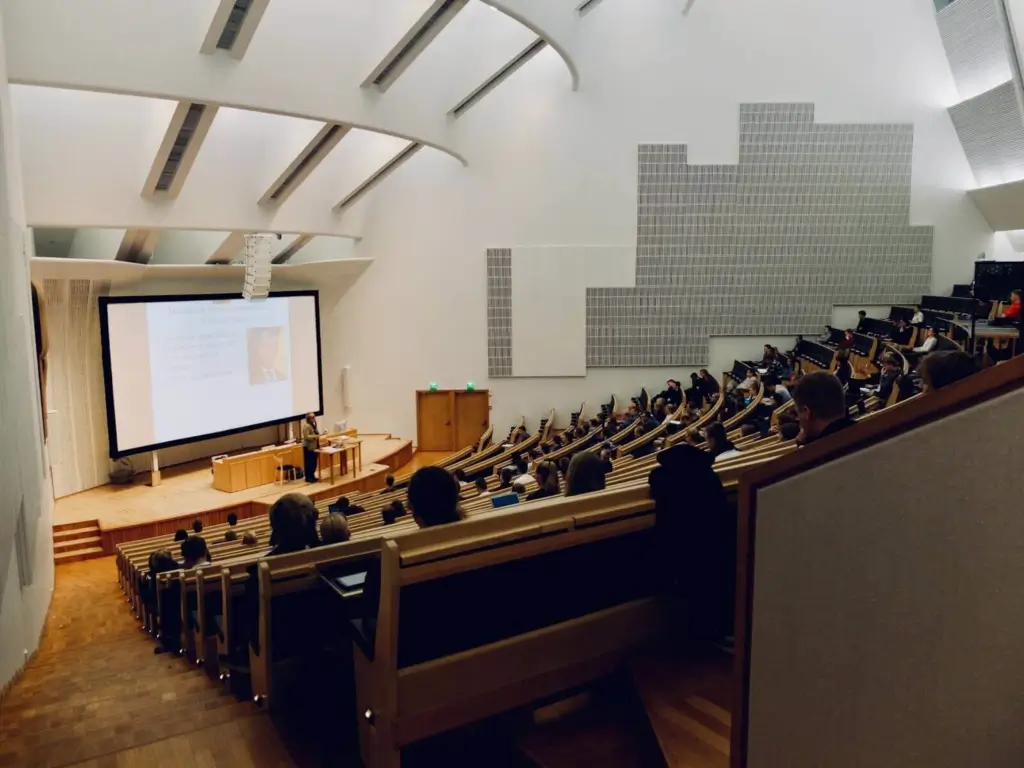 Large university lecture hall with tiered seating, where students listen to a presenter giving a lecture in front of a projected slide.