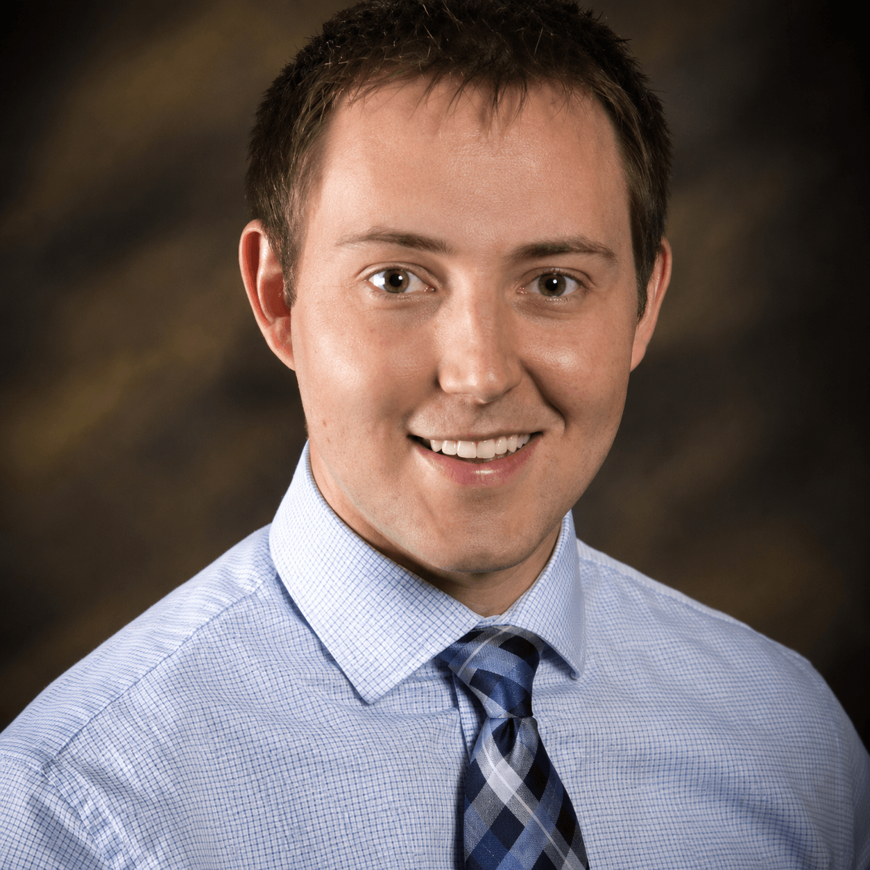 Professional headshot of a smiling man wearing a light blue dress shirt and patterned tie.